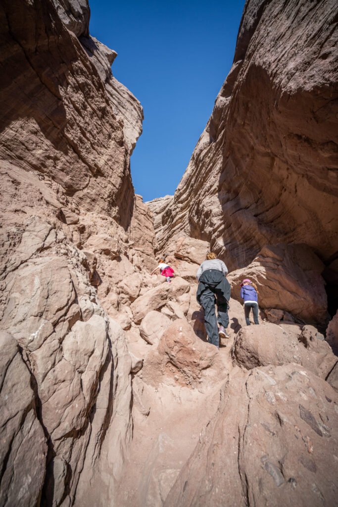 Hikers navigate a narrow canyon with towering sandstone walls and a vivid blue sky. The rugged landscape is a popular hiking trail in the desert southwest.