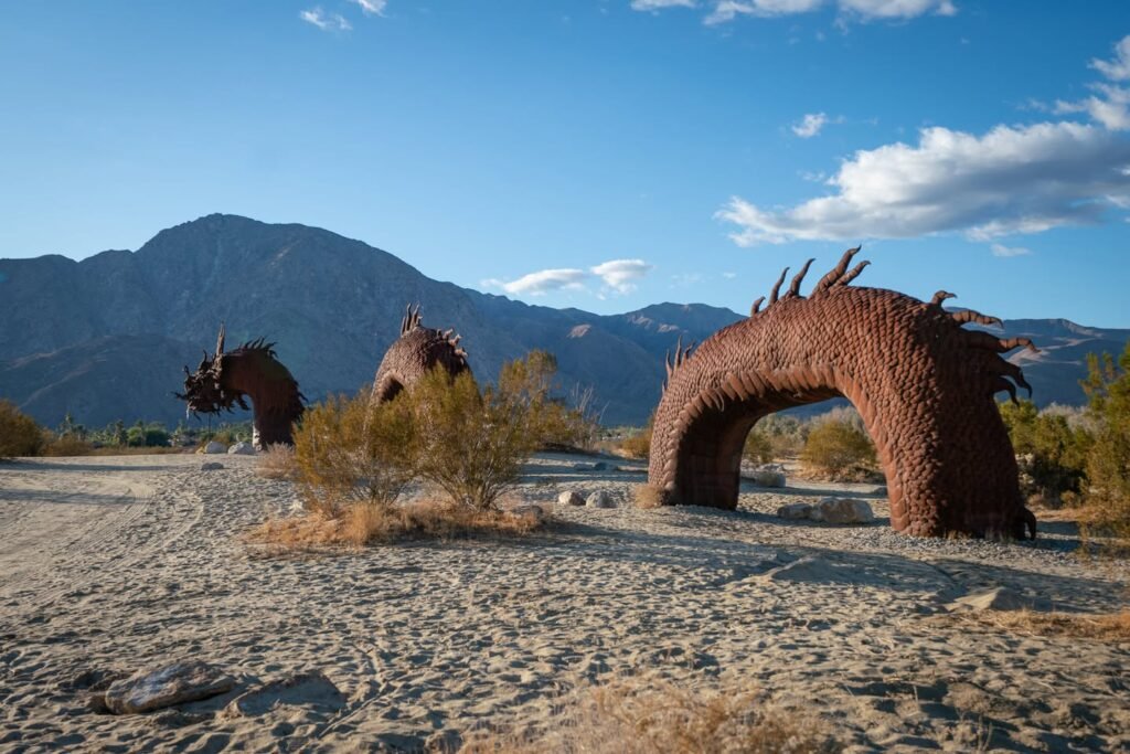 A wooden dragon sculpture in a desert landscape with mountains in the background. The scene is set under a clear blue sky.