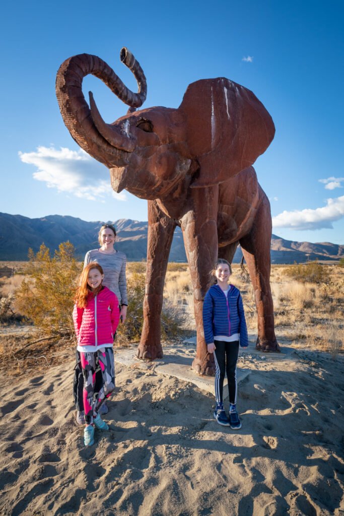 Visitors stand in front of a large rust-colored metal elephant sculpture in a desert landscape under a clear blue sky.