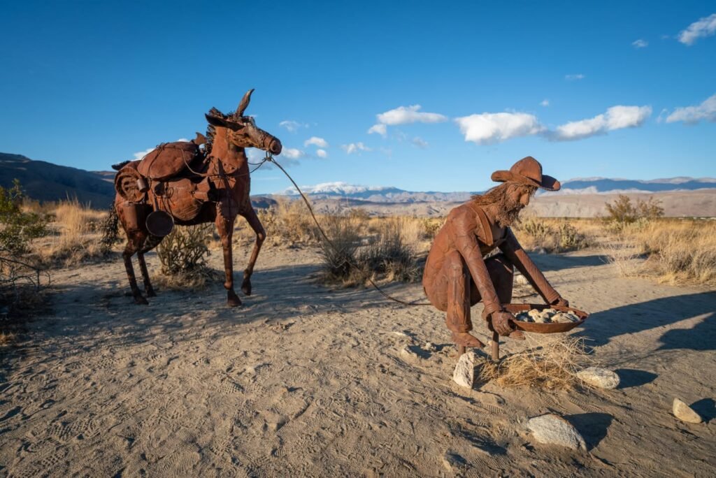 Outdoor sculptures depict a cowboy tending to a horse in a dry, open landscape. The figures are crafted from a textured, reddish material resembling rusted metal.