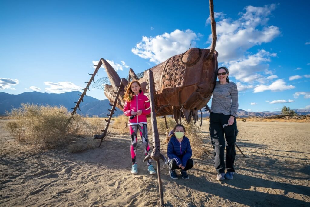 A family posing in front of an enormous rusted grasshopper sculpture in a desert environment under a bright blue sky.
