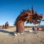 Ladder Canyon & Painted Canyon Two individuals standing near a striking dragon arch sculpture in a desert setting under a bright blue sky.