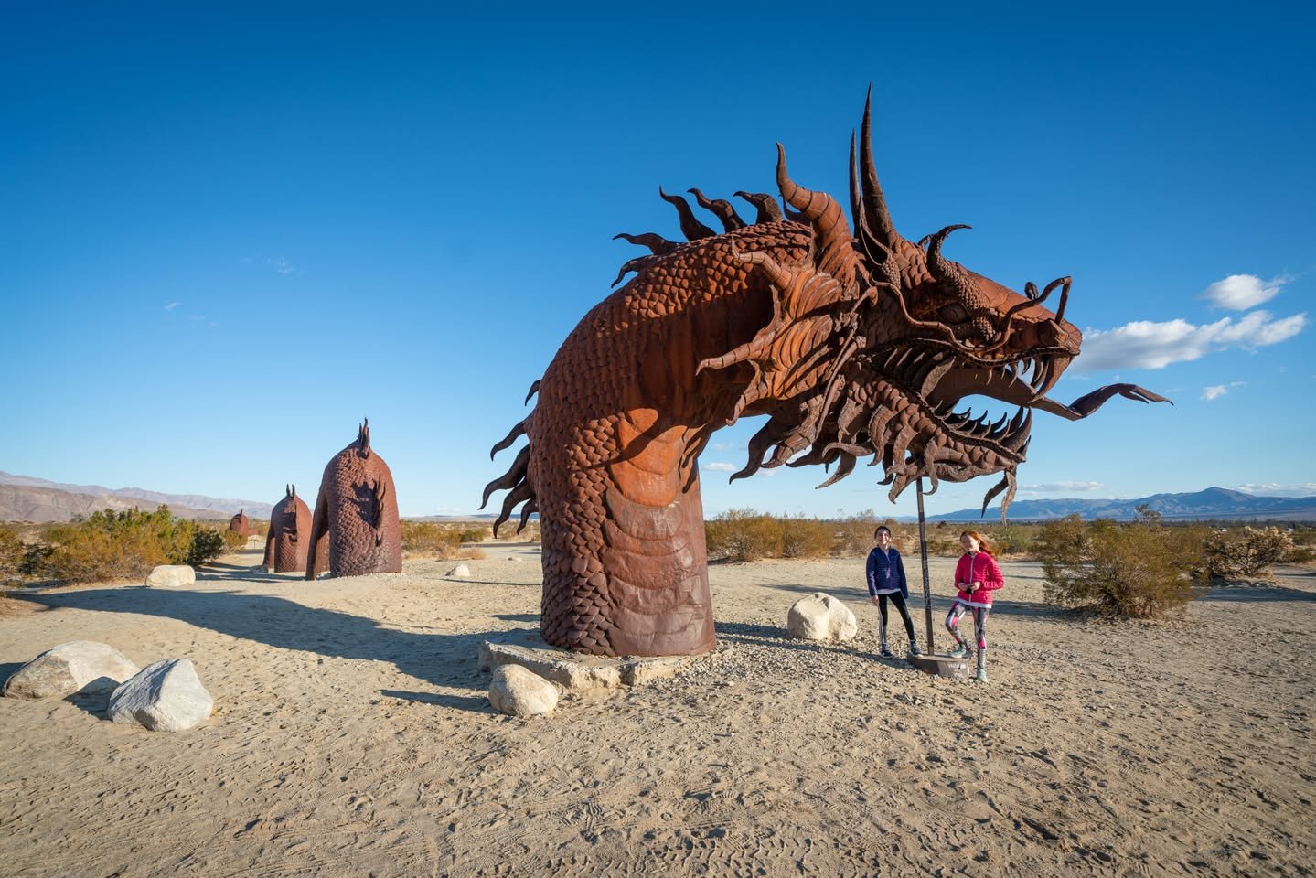 Anza-Borrego Sculptures Two individuals standing near a striking dragon arch sculpture in a desert setting under a bright blue sky.