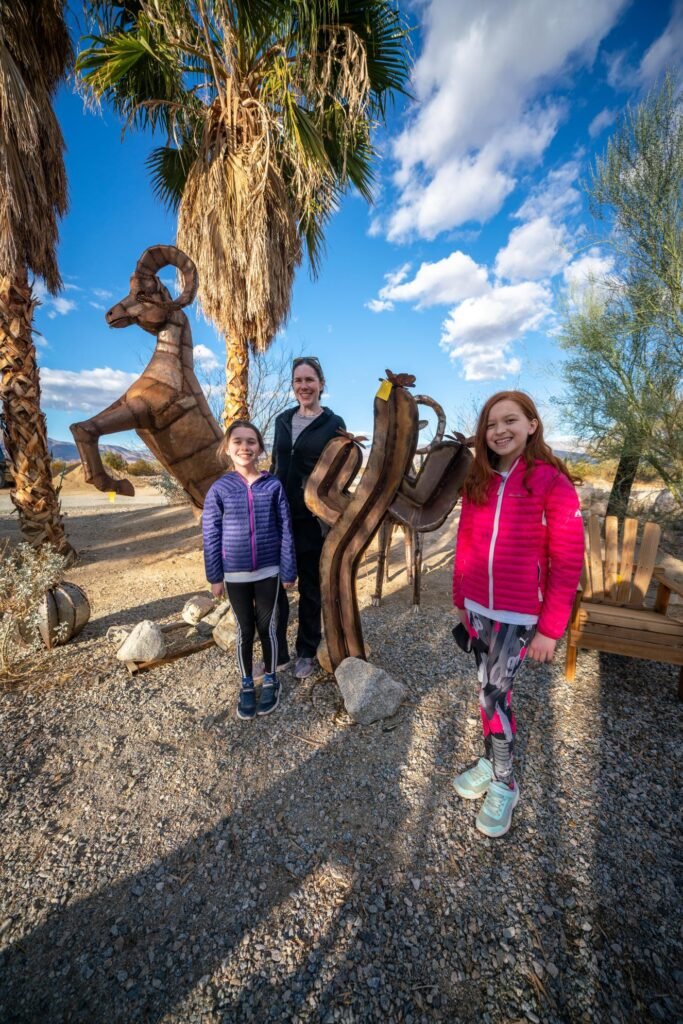 A family stands near a large metal ram sculpture surrounded by palm trees on a sunny day. The background features a bright blue sky with scattered clouds.
