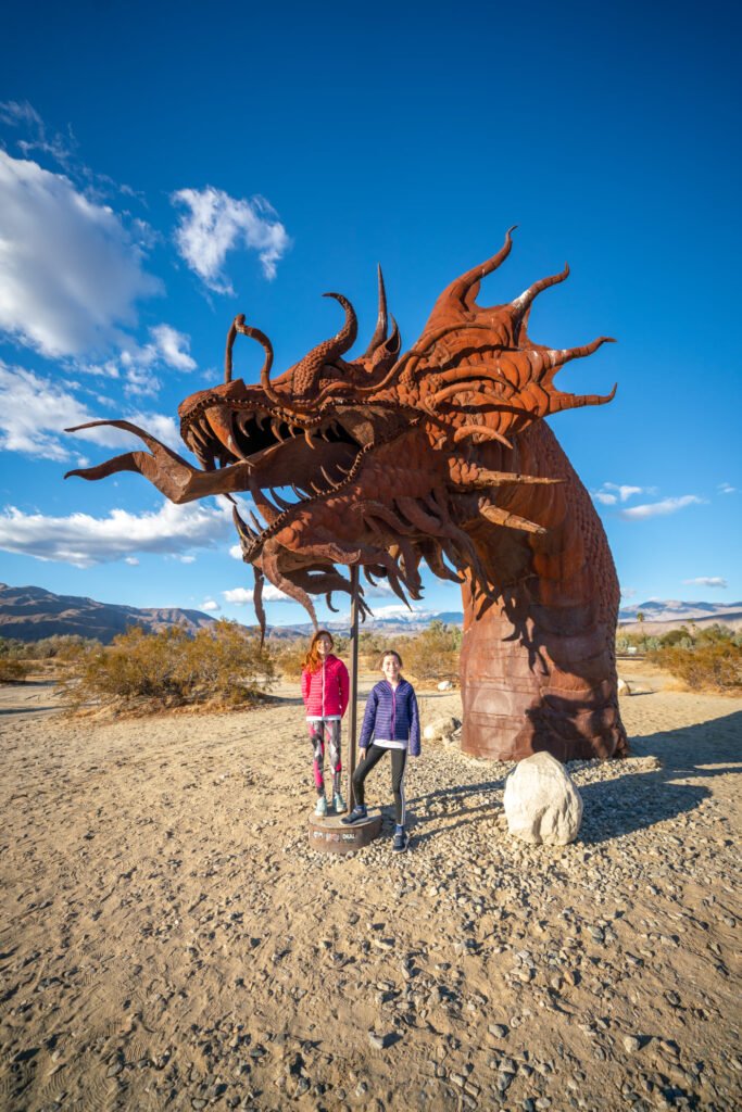 A rusted dragon sculpture on a desert road against a blue sky with clouds.