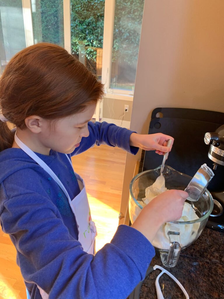 A child wearing a blue sweatshirt and apron stirring batter in a clear glass bowl using a stand mixer attachment.