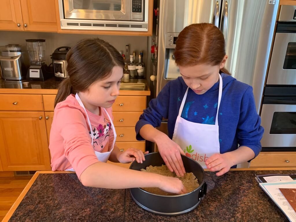 Two young individuals wearing aprons press a mixture into a cake pan on a kitchen counter. The scene includes stainless steel appliances and wooden cabinets.