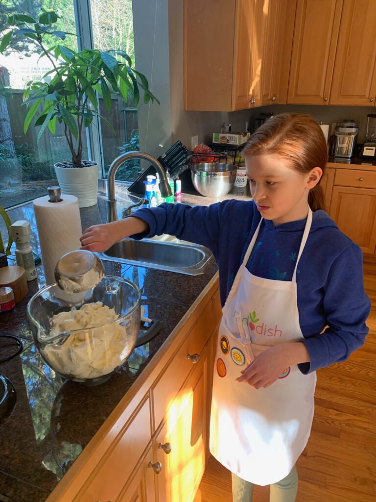 A young person with red hair wearing a white apron mixes flour in a glass bowl on a dark countertop during a cooking activity.