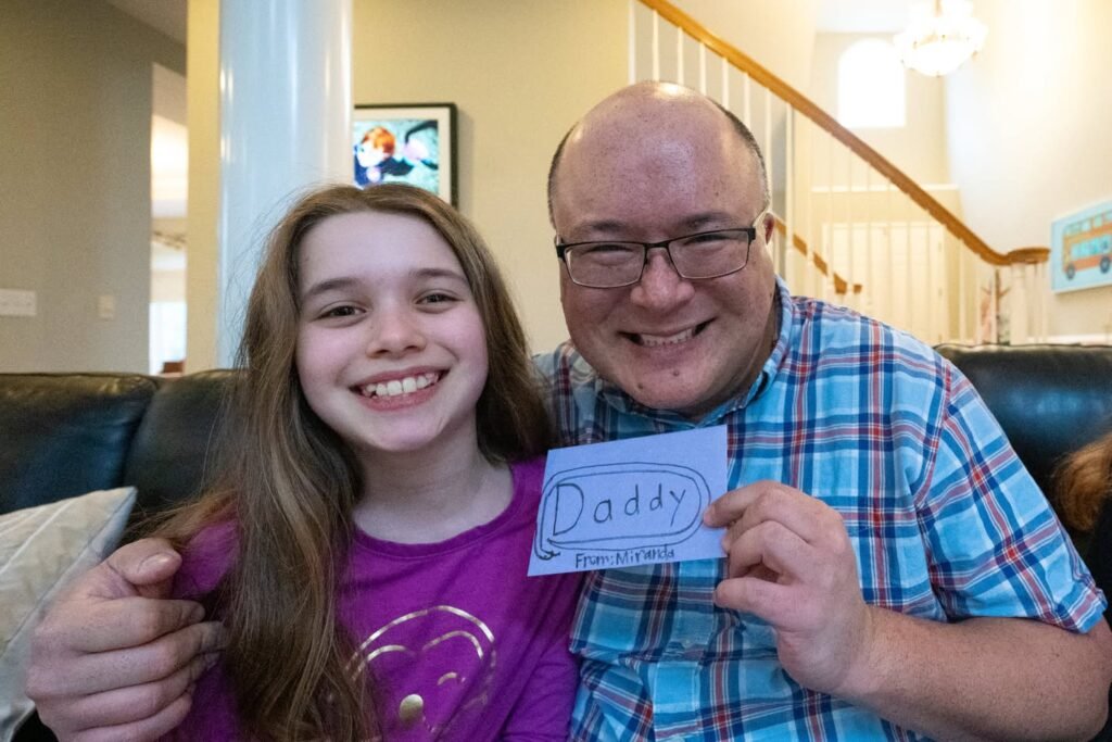Two people share a smile on a couch while holding up a blue card that reads Daddy. A cozy staircase and home decor complete the domestic background.