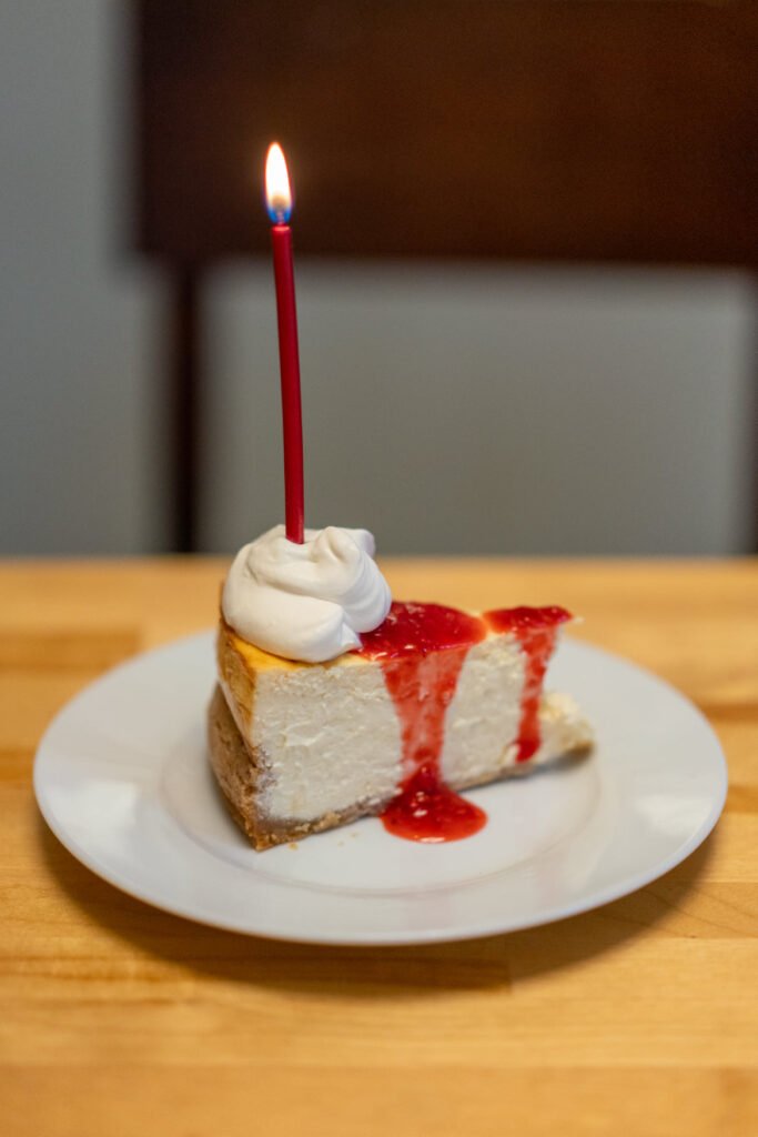 A slice of cheesecake topped with whipped cream, strawberry sauce, and a lit red candle. The dessert is served on a white plate.