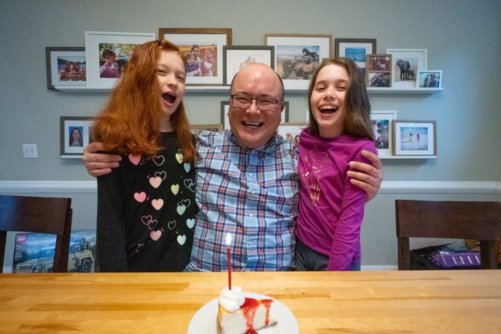 Three people smiling with a small cake on a wooden table in front of framed photos.
