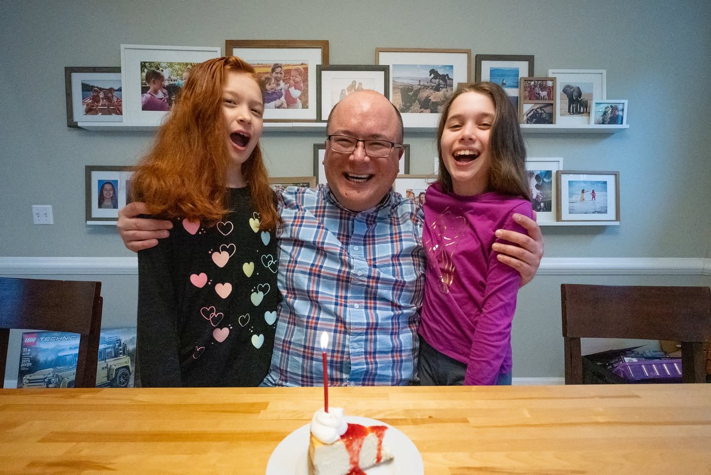 Three people smiling with a small cake on a wooden table in front of framed photos.