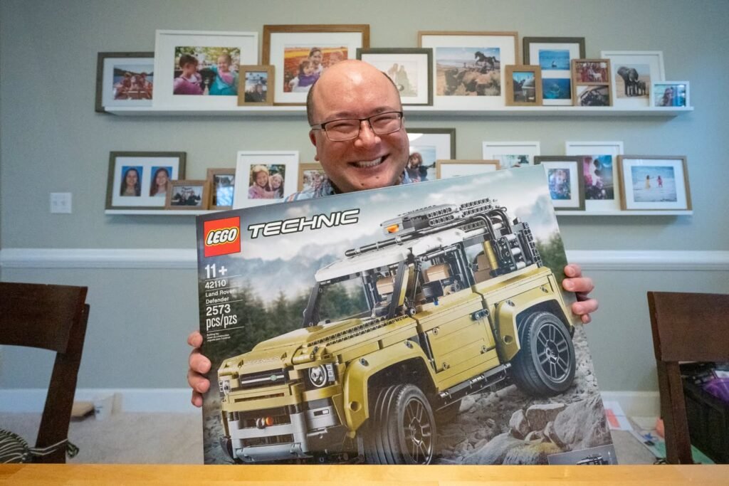A person holds a new Leg Technic Discovery set in front of a wall of family photos. The vintage vehicle design is displayed prominently on the set's box.