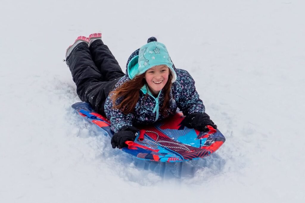 A person smiling while snoosle on a red and blue sled on a snowy slope.