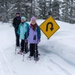 Snowy Valentine’s Three cross-country skiers wearing winter hats stand beside a snow-covered road next to a U-turn warning sign in a snowy forested area.