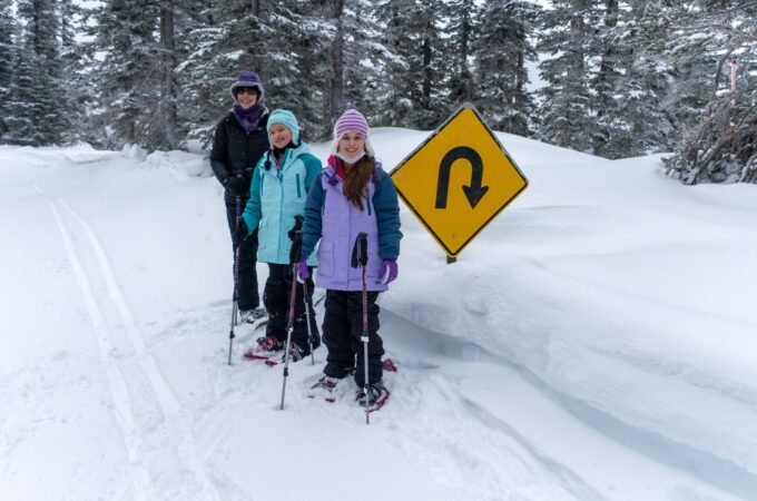 Three cross-country skiers wearing winter hats stand beside a snow-covered road next to a U-turn warning sign in a snowy forested area.