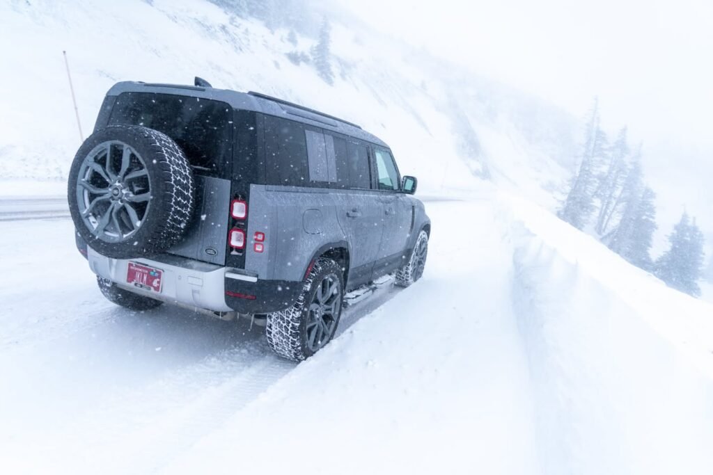 A rugged SUV equipped with chain-equipped tires drives through a snowy landscape. The vehicle features a rear-mounted spare tire against a backdrop of white mountains.