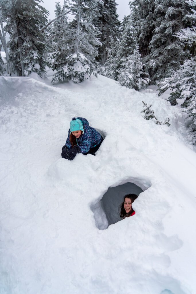 Two winter campers are nestled in deep snow next to a small tent in a snowy forest. Evergreen trees provide a wintry backdrop for this backcountry experience.