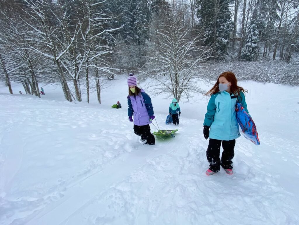 A small group of children play in a snowy forest setting. One child is seen pushing a sled through deep snow towards the camera.