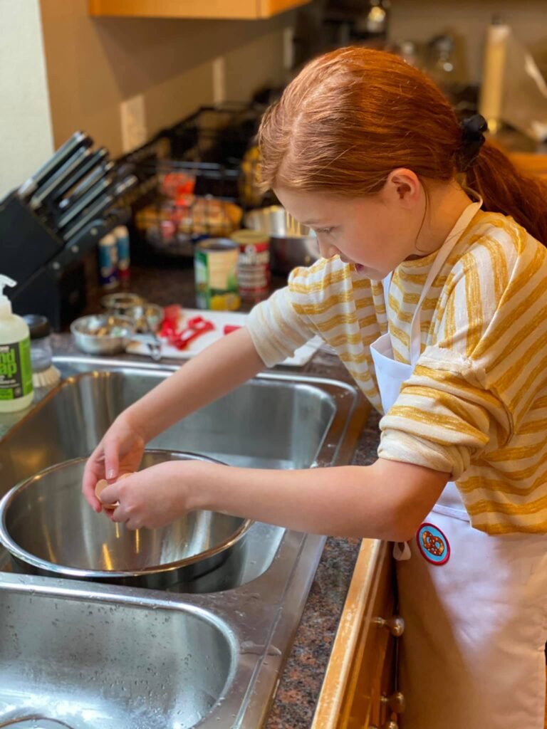 A small child cracks eggs into a bowl at the kitchen sink.