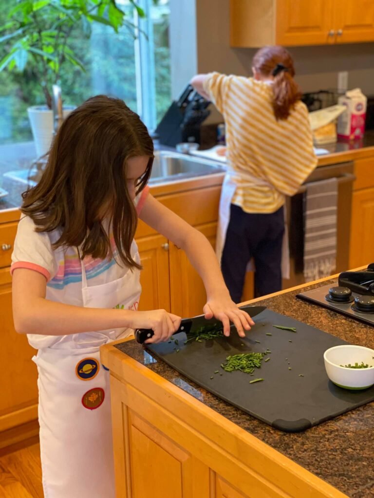 A young person chops fresh basil on a cutting board while another person works in the background.
