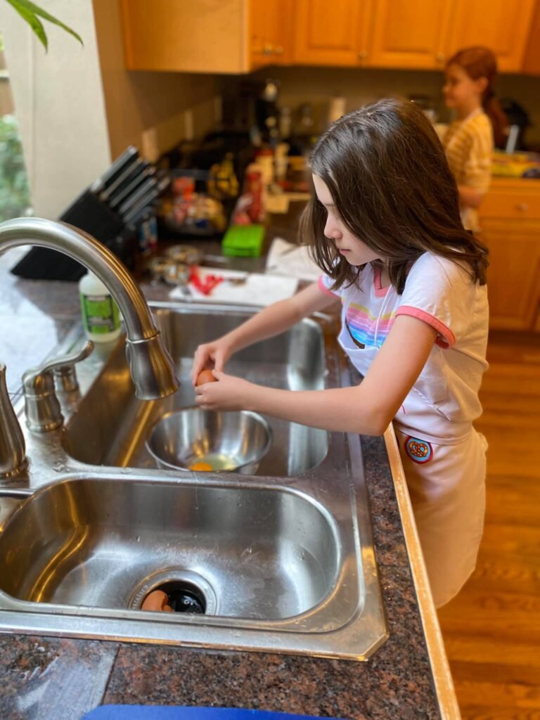 Children are washing dishes in a home kitchen sink, learning practical skills.