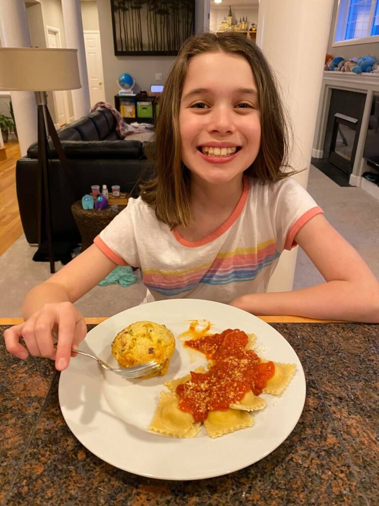 A child smiles while eating a plate of ravioli, mashed potatoes, and pepperoni in a cozy home kitchen.