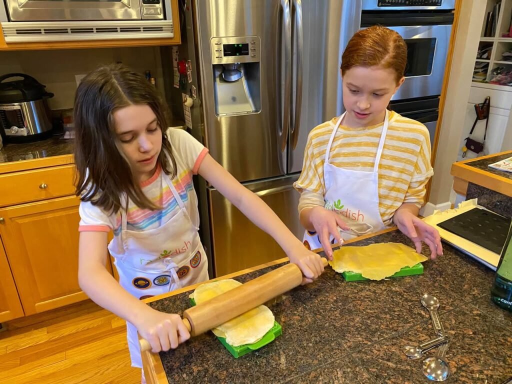 Two young people wearing aprons roll out green tortillas on a kitchen counter. A stainless steel refrigerator and wooden cabinets are visible in the background.