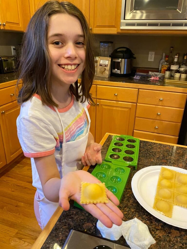 A child is smiling while making pasta using a green ravioli press on a kitchen counter. A plate of finished ravioli sits next to the cooking tools.