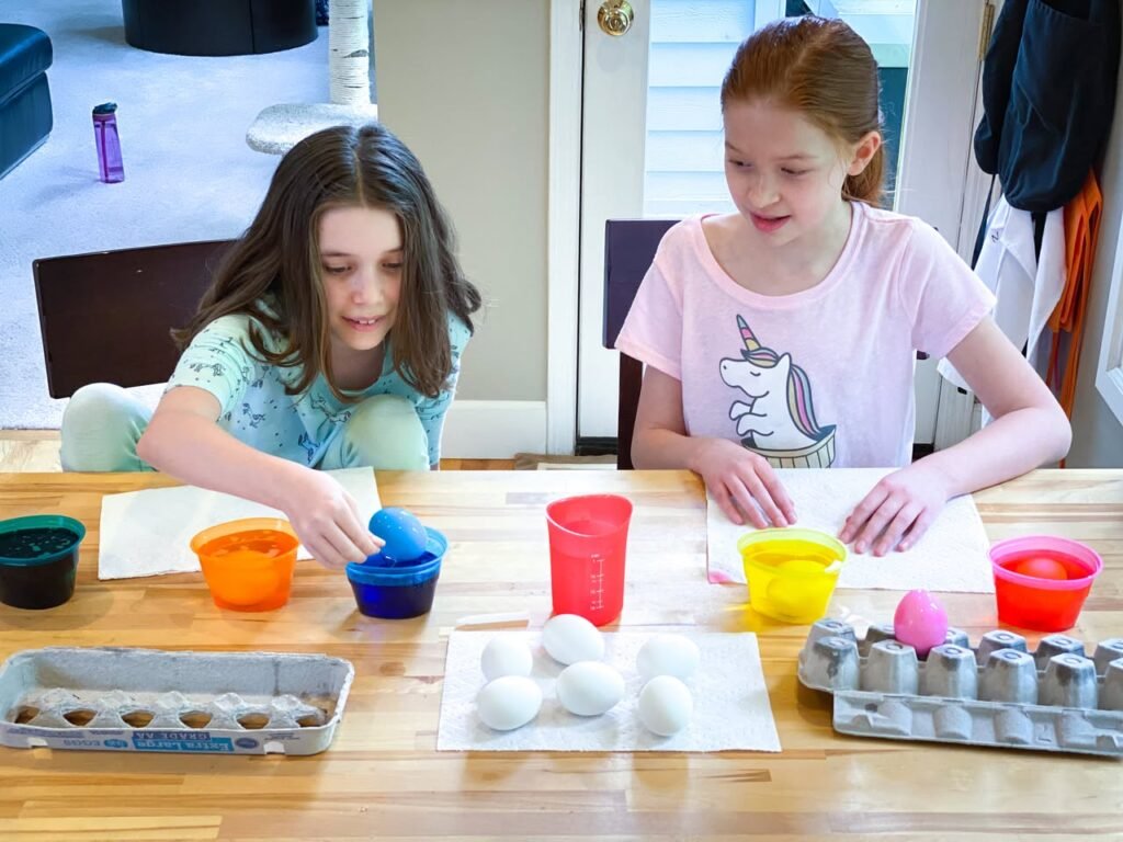 Two children dyeing Easter eggs at a table with colorful cups and trays. A view of the egg dyeing process and colorful setups.