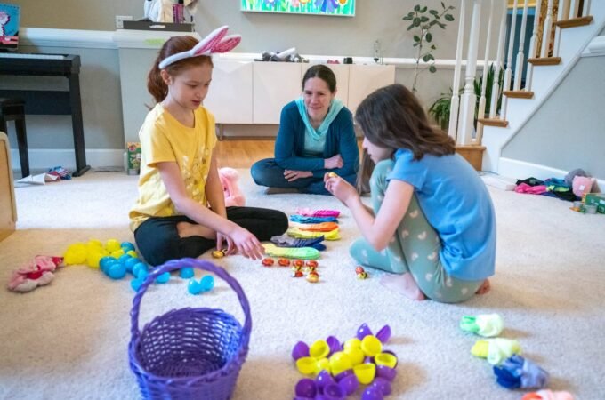Children and an adult collecting dyed Easter eggs in a cozy living room with a purple basket and colorful eggs on the floor.