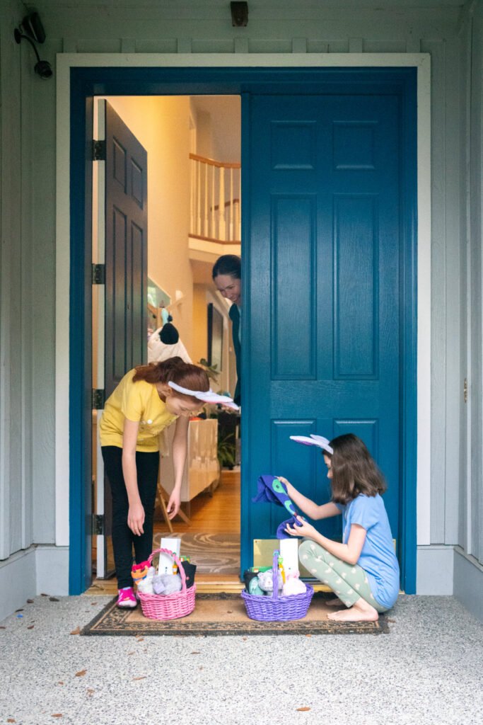 Children wearing bunny ears search for eggs in baskets at a blue front door.