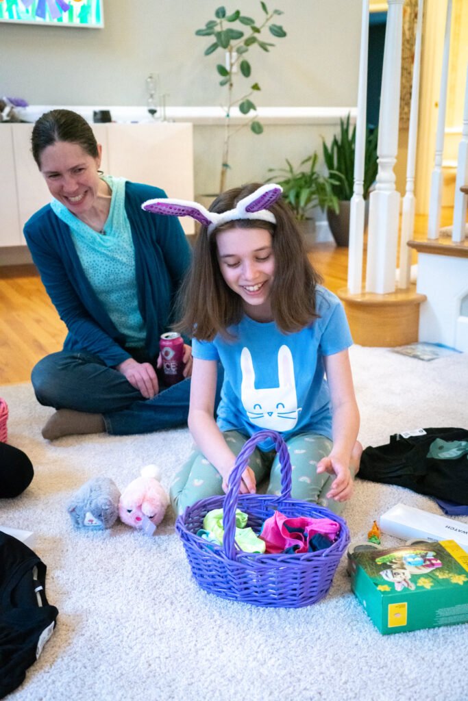 A girl wearing bunny ears sits on the floor with an Easter basket filled with eggs and toys while a woman watches with a smile.