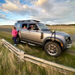 A silver SUV parked on a grassy roadside near a wooden fence with passengers looking out at the scenic landscape under a partly cloudy sky. The open terrain and vast skies highlight the beauty of rural travel.