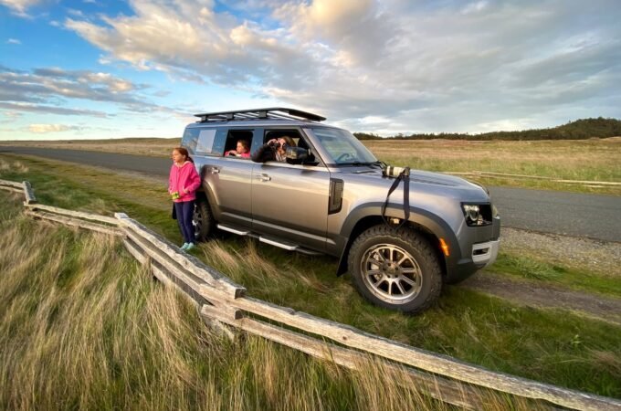 A silver SUV parked on a grassy roadside near a wooden fence with passengers looking out at the scenic landscape under a partly cloudy sky. The open terrain and vast skies highlight the beauty of rural travel.