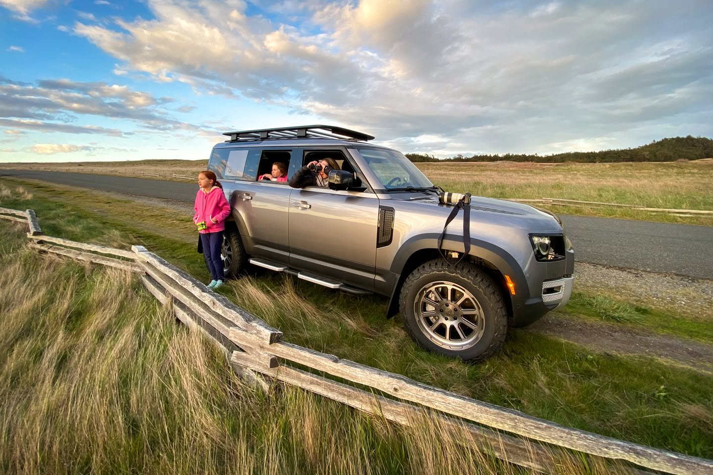 San Juan Safari A silver SUV parked on a grassy roadside near a wooden fence with passengers looking out at the scenic landscape under a partly cloudy sky. The open terrain and vast skies highlight the beauty of rural travel.