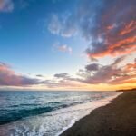 A serene beach scene in the Pacific Northwest at sunset, with waves rolling onto the shore. The sky is filled with pink and orange clouds.
