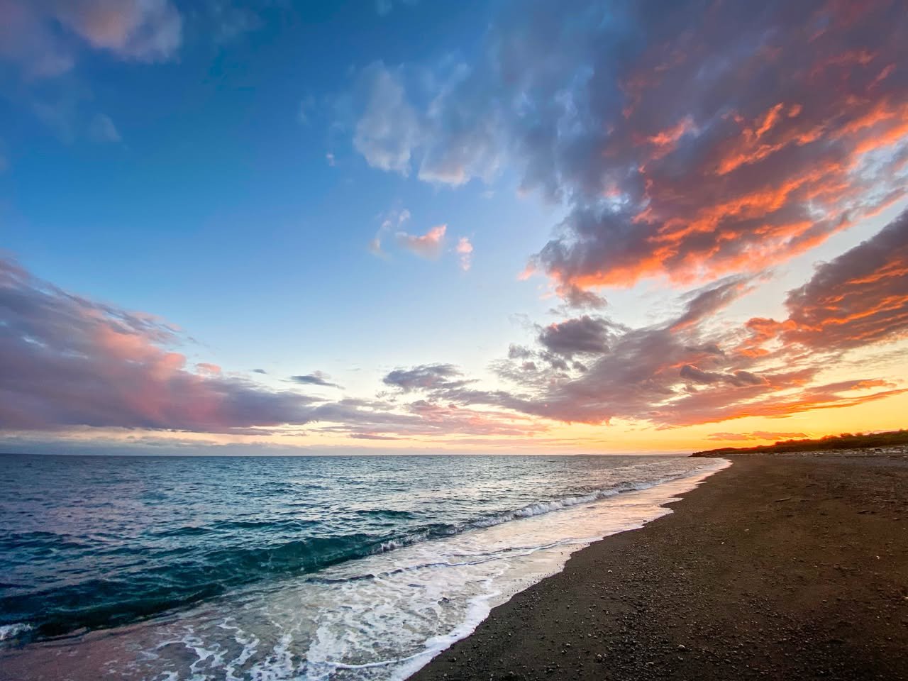 San Juan Sunsets & S’mores A serene beach scene in the Pacific Northwest at sunset, with waves rolling onto the shore. The sky is filled with pink and orange clouds.