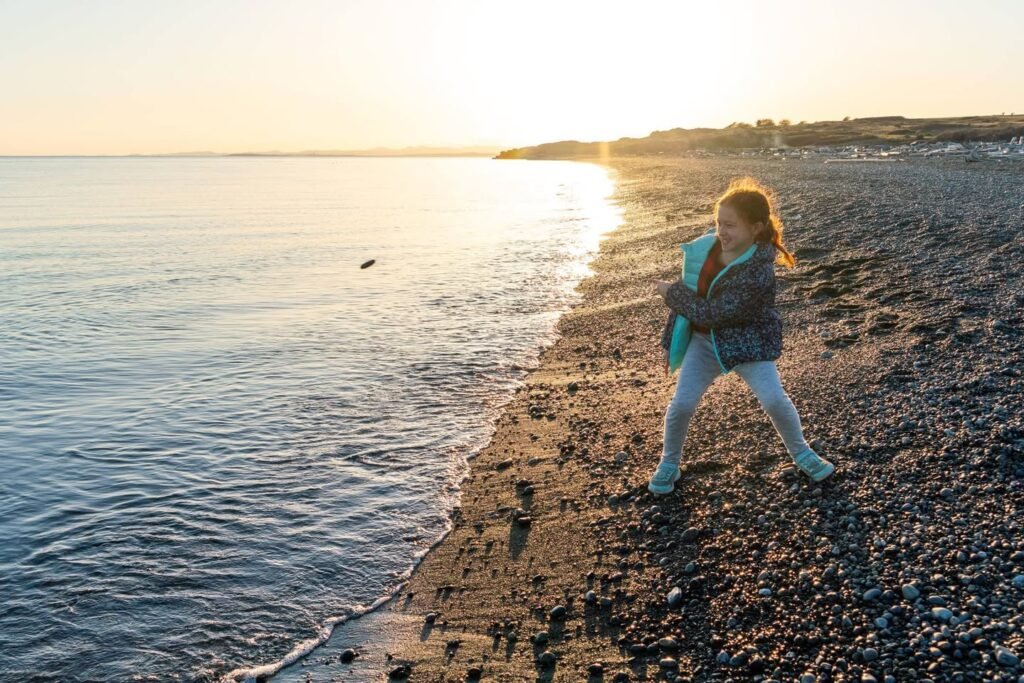 A person playing with a ball on a rocky shoreline at sunset. The scene features calm water meeting a pebble-filled beach.