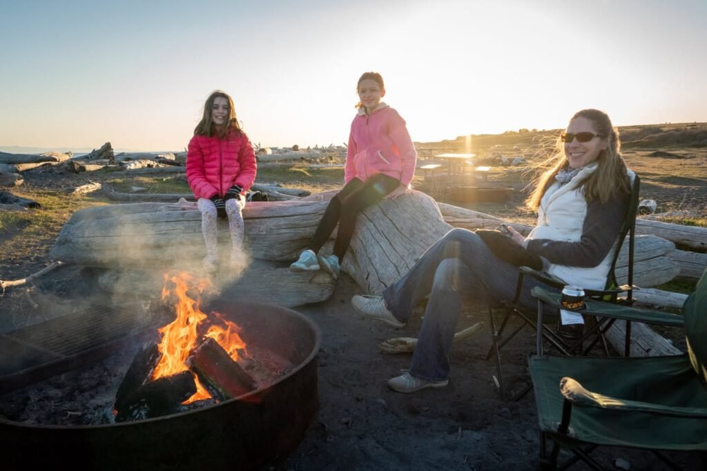 Three people relaxing by a beach fire pit at sunset. Close-up views of the flames and a person in a pink puffer jacket.