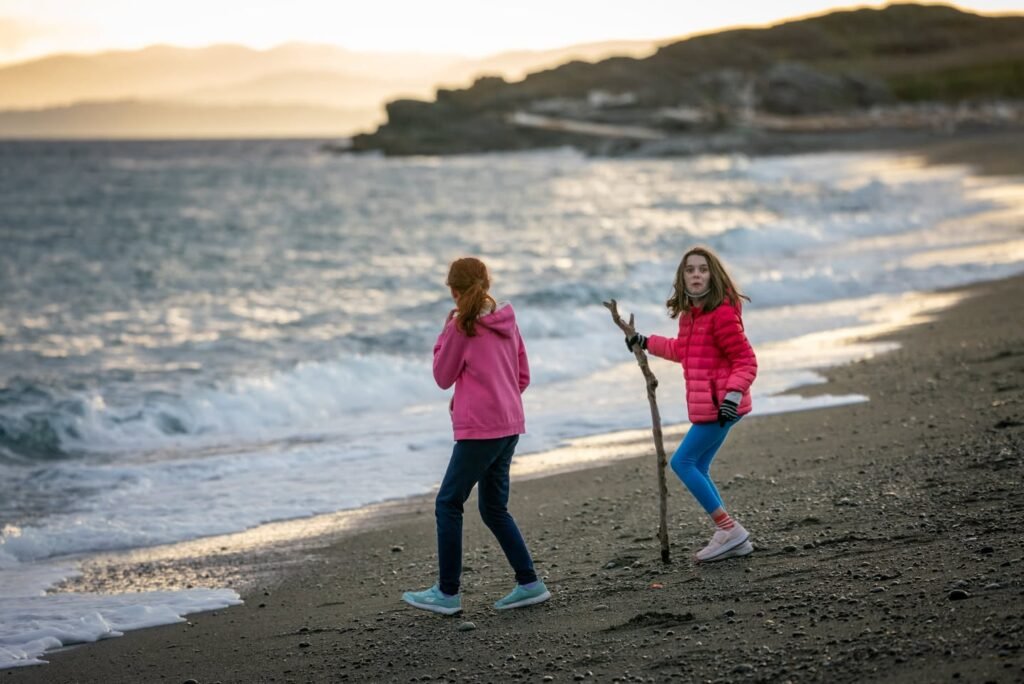 Two people walk along a sandy tide line during a scenic twilight walk. A rugged coastline features gentle rolling waves and warm golden light at the horizon.