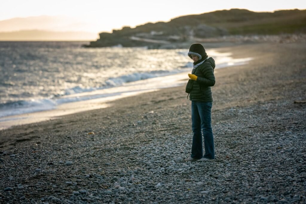 A young child bundled in warm clothing stands on a pebble beach, holding a yellow object against a backdrop of gentle waves and a distant headland.