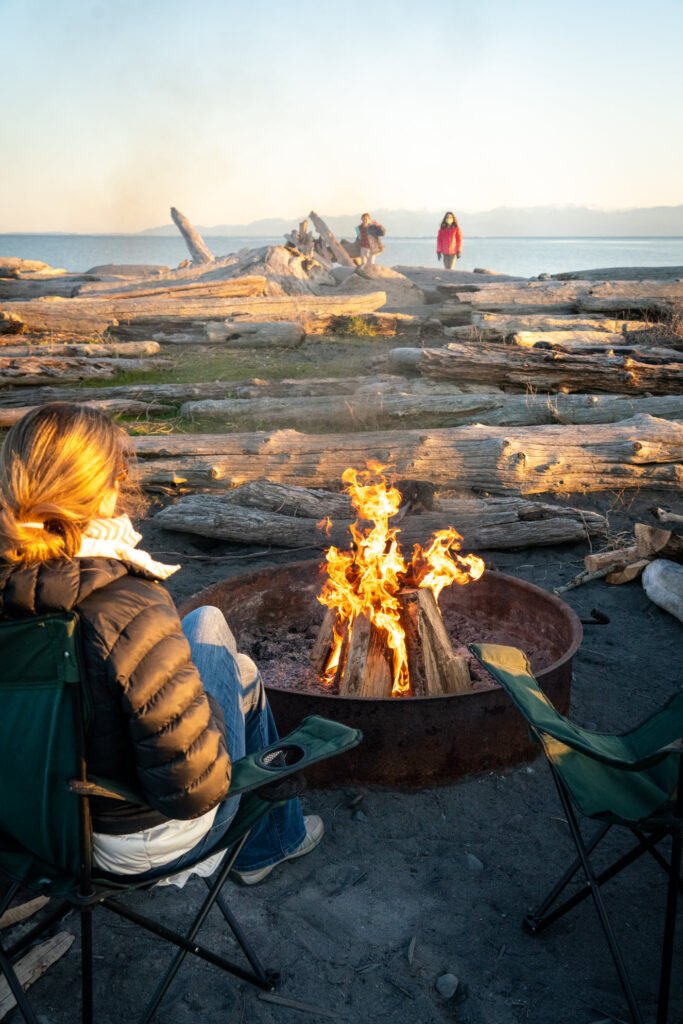 A campfire in a metal fire pit on a Pacific Northwest beach at sunset, with driftwood, ocean, and people enjoying the shore.