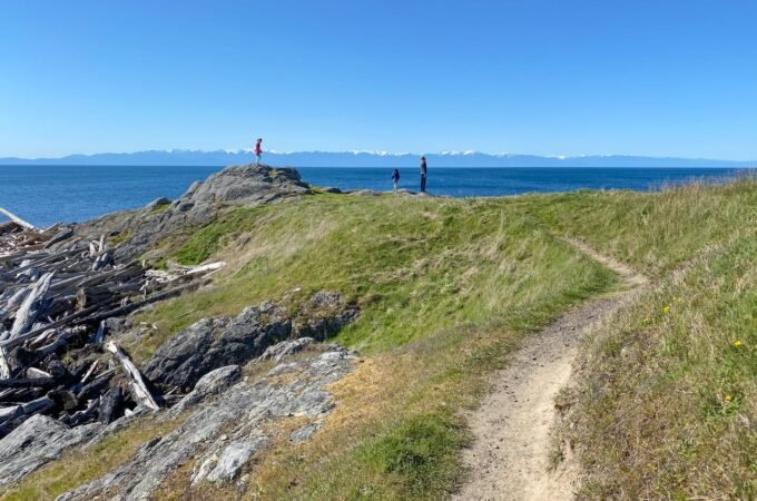 A dirt path winds along a coastal ridge with a view across the deep blue ocean to distant mountains under a clear blue sky.