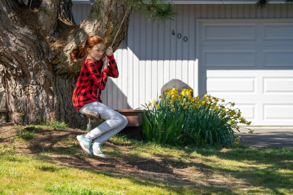 A person in a red jacket stands near a large tree in a front yard. Yellow flowers and a white garage door are visible in the background.