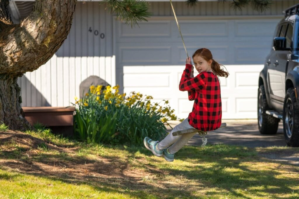 A person in a red plaid shirt swings from a tree in a sunlit driveway.