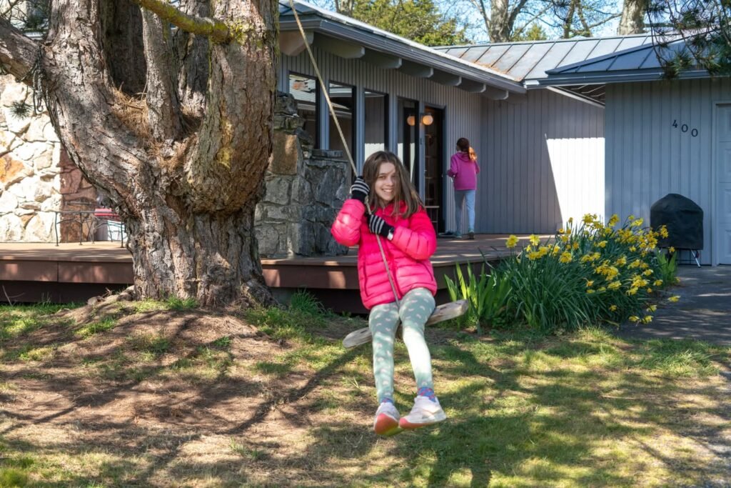 A person wearing a bright red jacket swings from a thick rope attached to a large tree in a backyard.
