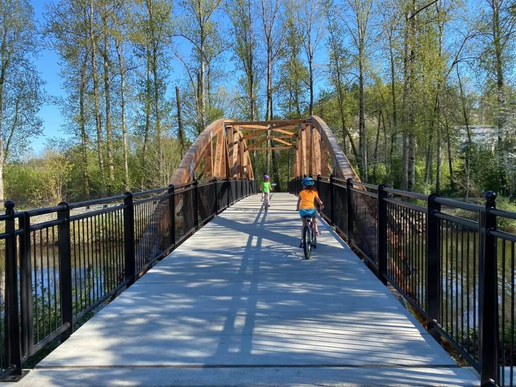 A wide boardwalk bridge with black railings crosses a small body of water surrounded by tall trees. A cyclist in an orange shirt is captured from behind crossing the wooden arch.