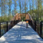 A wide boardwalk bridge with black railings crosses a small body of water surrounded by tall trees. A cyclist in an orange shirt is captured from behind crossing the wooden arch.