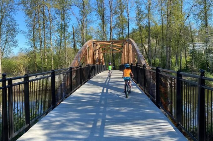 A wide boardwalk bridge with black railings crosses a small body of water surrounded by tall trees. A cyclist in an orange shirt is captured from behind crossing the wooden arch.